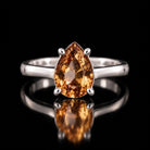 The same VooDooStone pear-cut solitaire ring turning golden amber under warm incandescent light, front view. Item 894782.